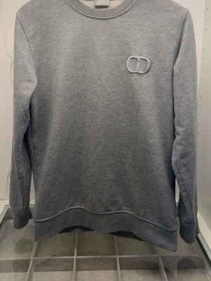 Grå sweatshirt med broderad logga - Säljer en stilren grå sweatshirt med rund hals och lång ärm. Tröjan har en broderad logga med två ringar på bröstet och är gjord i mjukt sweatshirtmaterial. Perfekt för en avslappnad look.