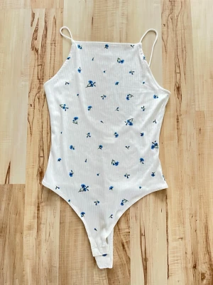 Vit ribbad body med blå blommor - Söt vit body med tunna axelband och ribbad struktur. Bodyn har ett blommigt mönster med små blå blommor och en hög halsringning fram samt djup rygg. Perfekt att styla till jeans eller kjol för en fräsch look.