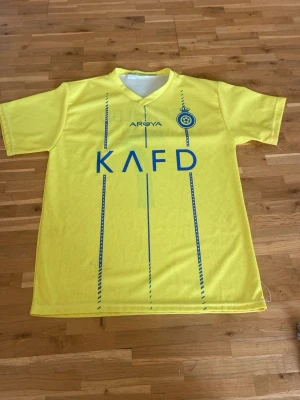 Gul Al Nassr Ronaldo matchtröja - Snygg gul matchtröja från Al Nassr med blå detaljer och tryck. Framsidan har klubbmärke, sponsorlogga och vertikala ränder. Baksidan har namnet 'Ronaldo' och nummer 7 i blått. Tillverkad i lätt och ventilerande material, perfekt för fotboll eller träning. Storlek s😁har små fläckar på framida ner