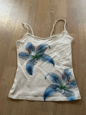 Vit linnetopp med blå blommor - Snygg vit linnetopp med tunna axelband och två stora blå blommor framtill. Toppen har en figurnära passform och är perfekt för sommar/vår dagar. Materialet känns mjukt och lätt, troligen bomull eller en bomullsblandning.