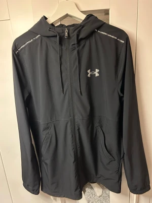 Svart vindjacka från Under Armour - Snygg svart vindjacka från Under Armour med dragkedja, huva och två fickor framtill. Jackan har logga på bröstet och text på axlarna. Tillverkad i lätt och vindtätt material, perfekt för blåsiga dagar. Storlek M