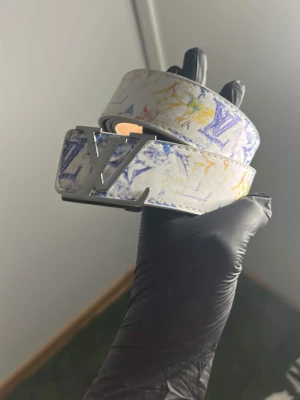 Vit bälte med färg - Snyggt vitt bälte från  med färgstänk i blått, gult och rött.. Bältet har ett metallspänne i silver med -logga och är gjort i skinn. Perfekt för att lyfta vilken outfit som helst.