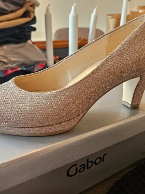 Glittriga pumps från Gabor - Säljer ett par eleganta platåpumps från Gabor i skimrande beige tyg med rundad tå och medelhög klack. Skorna har en klassisk siluett och är täckta med ett glittrigt material som ger extra wow-faktor. Perfekta för dig som vill sticka ut lite extra. Storlek 35.5 men passar 37. Endast provgått med dom inne men blev aldrig använda. Nypris 1350 kr.