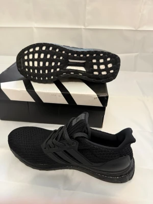 Svarta Adidas Ultra Boost sneakers - Säljer ett par helsvarta Adidas Ultra Boost sneakers med stickad ovandel och klassiska tre ränder på sidan. Skorna har en responsiv Boost-sula och svart yttersula med mönster för bra grepp. Perfekta för dig som gillar stilrena och bekväma sneakers.