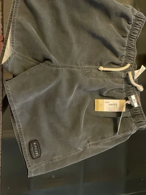Grå badbyxor från Rip Curl XL - Säljer ett par grå badbyxor från Rip Curl i storlek XL. De har en avslappnad passform, elastisk midja med vit dragsko och Rip Curl-logga på vänster ben. Materialet är snabbtorkande och insidan har meshfoder. Perfekta för stranden eller poolen!