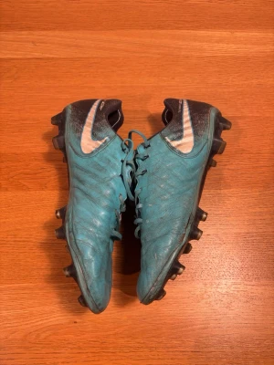 Nike blåa tiempo fotbollsskor - Säljer ett par blåa Nike fotbollsskor med svart häl och vit swoosh. Skorna har klassisk snörning och är tillverkade i syntetmaterial med struktur för bättre bollkontroll. Utrustade med fasta dobbar för spel på gräsplan.