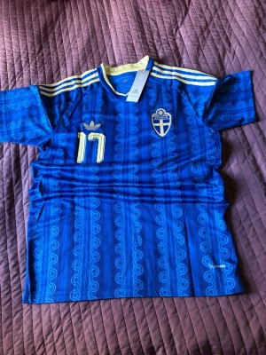 Blå Sverige fotbollströja Adidas - Säljer en blå fotbollströja från Adidas med Sveriges landslagsemblem, gula detaljer och nummer 17. På ryggen står det Gyökeres och stora siffror. Tröjan har korta ärmar, rund hals och ett mönster i tyget. Perfekt för match eller träning.