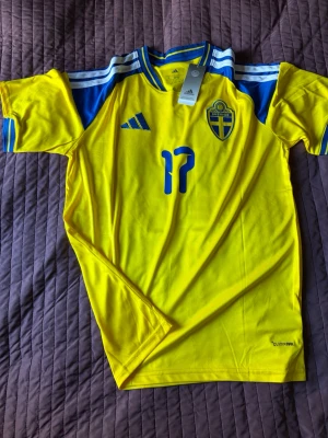 Sverige fotbollströja Adidas gul - Säljer en officiell gul Sverige fotbollströja från Adidas med blå detaljer, nummer 17 och namnet Gyökeres på ryggen. Tröjan har blå och vita ränder på axlarna, broderad landslagssköld och är tillverkad i lätt funktionsmaterial (Climalite). Perfekt för match eller träning!