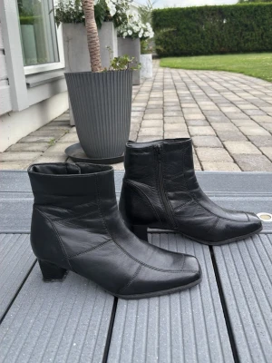 Svarta stövletter i äkta skinn med klack i strl 39 - Snygga svarta stövletter i äkta skinn med markerade sömmar och dragkedja på sidan. Modellen har en fyrkantig tå och stabil låg klack som ger en cool och stilren look. Perfekta till jeans eller kjol för en trendig outfit.