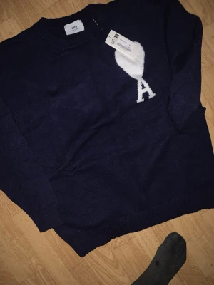 Mörkblå stickad tröja från Ami - Mörkblå stickad tröja från Ami med rund halsringning och ribbade muddar. På bröstet finns ett vitt hjärta med bokstaven 'A' i samma färg. Tröjan har en klassisk passform och är tillverkad i mjukt material, perfekt för lager på lager.