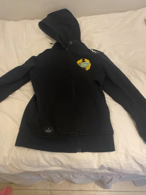 Svart hoodie från Craft med tryck - Svart hoodie från Craft med dragkedja och huva. På bröstet finns ett färgglatt klubbmärke i gult, grönt och vitt. Hoodien har snörning i huvan och ribbade muddar. Perfekt för dig som gillar sportig stil.
