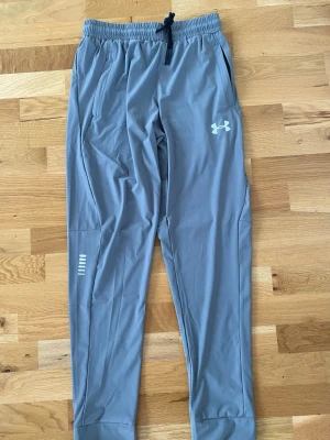 Grå mjukisbyxor från Under Armour  st. M men små i storleken - Snygga grå mjukisbyxor från Under Armour med resår i midjan och dragsko. Byxorna har två sidofickor, smal passform och logga på vänster lår. Tillverkade i ett mjukt och stretchigt material som passar perfekt för chill eller träning.