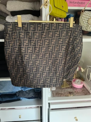 Beige och svart kortkjol - Säljer en beige och svart kortkjol från Fe med det klassiska F…-monogrammet över hela kjolen. Kjolen har en rak passform och är tillverkad i ett slätt material som känns lyxigt. Perfekt för dig som vill ha en statement piece i garderoben. Resår i midjan