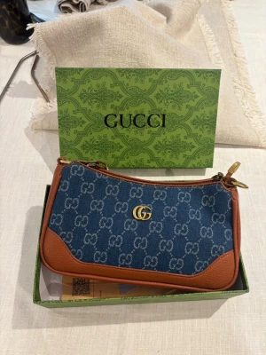 Gucci blå kuvertväska i denim - Snygg kuvertväska från Gucci i blå denim med GG-monogram och bruna läderdetaljer. Väskan har guldfärgad logga framtill och dragkedja upptill. Perfekt storlek för det viktigaste och levereras i originalförpackning med tillbehör.