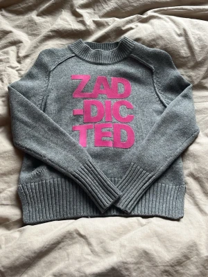 Grå stickad tröja Zadig & Voltaire - Grå stickad tröja från Zadig & Voltaire med stora rosa bokstäver 'ZADDICTED' framtill. Tröjan har rund hals, långa ärmar och breda ribbade muddar vid ärmslut och nederkant. Strl xxs/xs