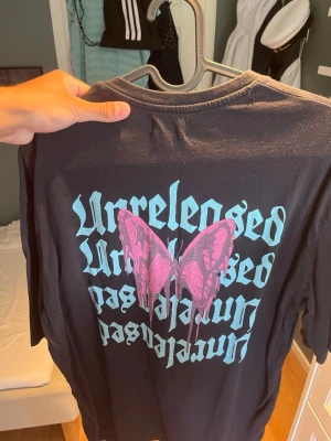 Svart t-shirt med fjäril från Yourturn - Svart t-shirt från Yourturn med trycket 'Unreleased' i turkos text på bröstet och stor rosa fjäril samt text på ryggen. Klassisk rund hals och korta ärmar.