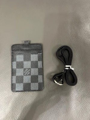  korthållare i svart  - Snygg korthållare från  i svart och grått rutmönster, tillverkad i canvas med läderdetaljer. Kommer med en svart rem som kan fästas i korthållaren. Perfekt för kort och ID med en transparent ficka på baksidan.