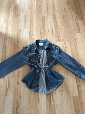 Blå jeansjacka från ONLY, XL - Snygg blå jeansjacka från ONLY i klassisk denim med knappar framtill, två bröstfickor och ett knytskärp i midjan. Jackan har lång ärm och en figurnära passform som ger en trendig siluett. Perfekt för att lyfta din outfit med en cool touch.