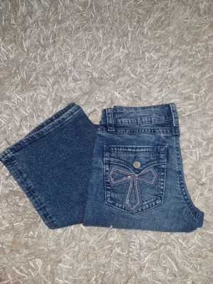 Blå jeans med paljetkorts - Snygga blå bootcut low waist jeans och en unik bakficka som har ett broderad kors i paljetter. Perfekta för dig som vill ha något extra detaljerat på dina jeans. från gina young, 170