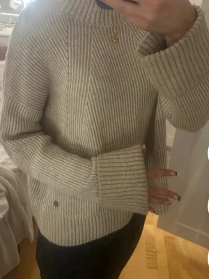 Beige ribbstickad tröja - Säljer en beige ribbstickad tröja med breda ärmar och hög hals. Tröjan har ett snyggt mönster med olika riktningar på stickningen och är perfekt för dig som gillar en avslappnad men ändå stilren look.