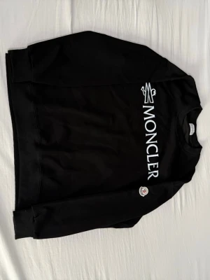 Svart Moncler sweatshirt  - Snygg svart sweatshirt från Moncler med stor vit logga tryckt på bröstet och Moncler-märke på ärmen. Tröjan har rund halsringning och ribbade muddar vid ärmslut och nederkant. Perfekt för en clean och trendig look. Helt ny och aldrig använd.