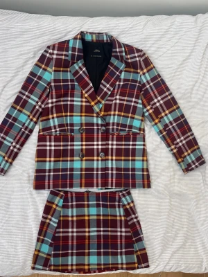 Urban Outfitters Rutig Kavaj och Kjol Set - Snygg dubbelknäppt kavaj med rutigt mönster i vinrött, blått, gult och vitt. Klassisk krage och raka ärmar. Materialet känns som polyester och bomull. Perfekt för dig som vill sticka ut med färg och mönster.