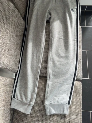 Gråa mjukisbyxor från Adidas - Snygga gråa mjukisbyxor från Adidas med svarta ränder längs sidorna och ribbade muddar vid bensluten. Byxorna har elastisk midja och är tillverkade i mjukt material, perfekta för chill dagar. Adidas-logga på vänster lår.