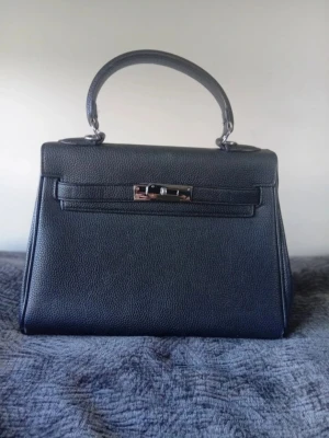Svart klassisk handväska i läder - Stilren svart handväska i strukturerat läder med silverfärgade metalldetaljer och ett topphandtag. Väskan har en elegant, boxig form och klassiskt vridlås framtill. Perfekt för dig som gillar tidlös design och vill ha en väska som sticker ut.