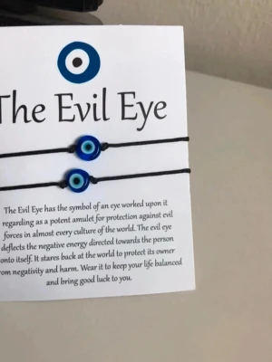 Evil Eye armband med pärla - Två svarta armband med blå Evil Eye-pärlor. Pärlorna har ett ögonmotiv i blått, vitt och svart och sitter på tunna svarta snören med sin citat broschyr och betydelse. Perfekt accessoar för dig som gillar symboliska smycken 