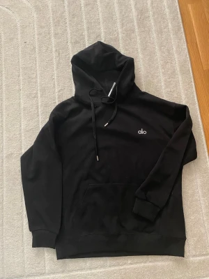 Svart hoodie från Alo - Snygg svart hoodie från Alo med minimalistisk vit logga på bröstet. Hoodien har en stor huva med dragsko, magficka och ribbade muddar vid ärmar och nederkant. Tillverkad i mjukt material som känns skönt mot huden. Perfekt för en avslappnad och trendig look.