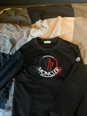 Svart Moncler!!!! - Svart sweatshirt från Moncler med stor broderad logga i vitt, rött och svart på bröstet. Tröjan har rund halsringning och Moncler-märke på ärmen. Perfekt för dig som gillar streetwear och exklusiva märken.