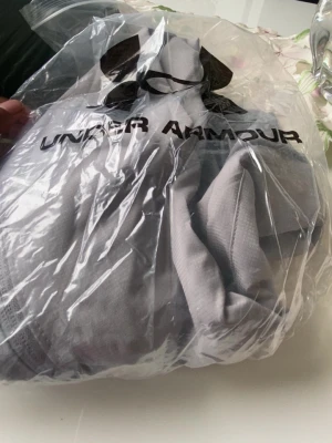 Ljusgrå vindjacka från Under Armour - Snygg ljusgrå vindjacka från Under Armour med huva och dragkedja framtill. Jackan har två sidofickor och är tillverkad i ett lätt, vindtätt material som passar perfekt för blåsiga dagar. Stort logotryck på bröstet ger en sportig look.storlek S/M
