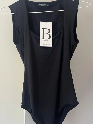 Svart basic body från Stradivarius - En svart, ärmlös body från Stradivarius Basics med rund halsringning. Tillverkad i mjukt och stretchigt material som sitter tight mot kroppen. Perfekt att styla med jeans eller kjol för en clean och enkel look.