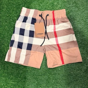 Rutig shorts från Burberry - Säljer ett par snygga rutig shorts från Burberry med klassiskt beige, svart, vit och röd mönster. Shortsen har snörning i midjan och är perfekta för sommaren. De har en loose passform och är tillverkade i ett lätt syntetmaterial.