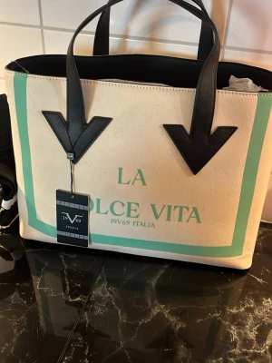 La Dolce Vita shoppingväska 19V69 - Trendig shoppingväska från 19V69 Italia i beige canvas med mintgröna detaljer och texten 'La Dolce Vita' framtill. Väskan har svarta handtag i konstläder och en rektangulär form, perfekt för att bära allt du behöver. Snygg och rymlig design med italiensk känsla. Slutsåld!