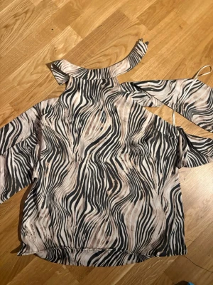 Zebramönstrad offshoulder blus - Snygg blus med zebramönster i svart, beige och vitt. Offshoulder-modell med hög hals och långa ärmar. Tunn och luftig i materialet, perfekt för att sticka ut i mängden. Trendig design som passar till många olika stilar.