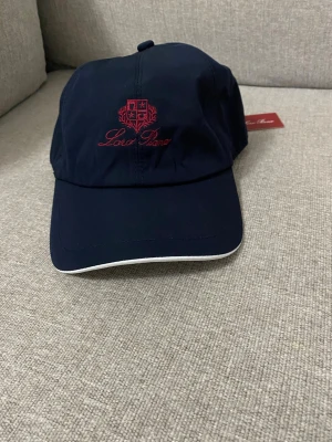 Mörkblå keps från Lord Piana - Snygg mörkblå keps från Lord Piana med röd broderad logga och text framtill. Kepsen har böjd skärm med vit kant och justerbar rem bak. Tillverkad i ett lätt och bekvämt material som passar perfekt till streetwear eller casual outfits.