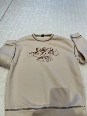 Beige sweatshirt med tryck Angel Baby - Beige sweatshirt med brunt tryck av änglar och texten 'ANGEL BABY' på bröstet. Tröjan har rund halsringning, ribbade muddar och synliga sömmar i kontrastfärg. Perfekt för en avslappnad och trendig look.