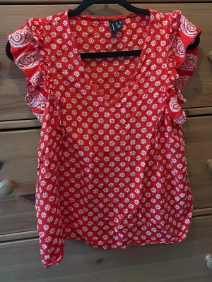 Röd blommig topp från Vero Moda - Superfin röd topp från Vero Moda med vita blommor, volangdetaljer på axlarna och v-ringning. Perfekt för dig som vill ha en färgglad och söt look. Luftigt material som känns skönt mot huden.