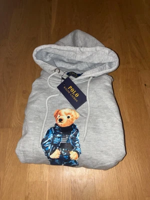 Grå hoodie Polo Ralph Lauren björn - Snygg grå hoodie från Polo Ralph Lauren med den ikoniska björnen på bröstet, klädd i blå jacka och jeans. Hoodien har dragsko i huvan och ribbade muddar vid ärmar och nederkant. Perfekt för dig som gillar streetwear och vill ha något unikt.