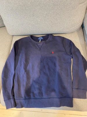 Mörkblå sweatshirt från Polo Ralph Lauren - Mörkblå sweatshirt från Polo Ralph Lauren med klassisk rund halsringning och diskret röd logobrodyr på bröstet. Tröjan har långa ärmar och är gjord i mjukt bomullsmaterial. Perfekt för en avslappnad och stilren look.