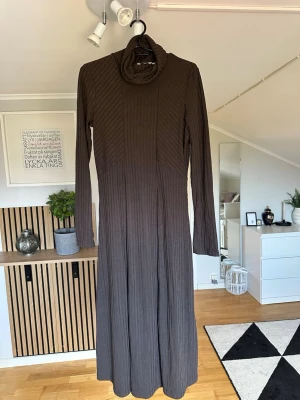 Brun långklänning från Indiska - Mysig brun långklänning från Indiska med polokrage och ribbat mönster. Klänningen har långa ärmar och är tillverkad i ett mjukt material som ger en skön passform. Perfekt för dig som gillar en enkel men stilren look.