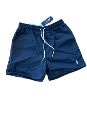 Svarta badbyxor från Polo Ralph Lauren - Snygga marinblå badbyxor från Polo Ralph Lauren med vit dragsko i midjan och klassisk logga broderad på benet. Tillverkade i snabbtorkande material och har en bakficka med lock. Perfekta för stranden eller poolen.