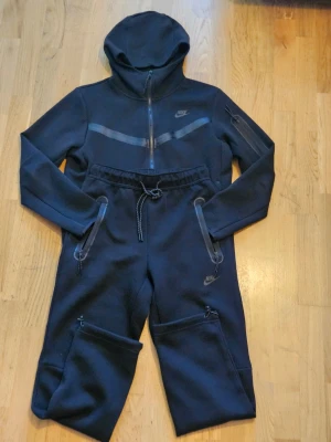 Svart Nike Tech Fleece tracksuit - Säljer en svart Nike Tech Fleece hoodie med dragkedja och huva. Hoodien har diskreta reflexdetaljer över bröstet och en ficka med dragkedja på ärmen. Tillverkad i mjukt och bekvämt fleecematerial, perfekt för en sportig och avslappnad stil.
