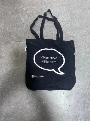 Svart tygkasse Göteborgs Stad - Svart shoppingväska i tyg med dubbla handtag. Ena sidan har ett vitt pratbubblemotiv med texten 'Vilken skola väljer du?' och Göteborgs Stad-logga. Andra sidan har flera vita logotyper från olika gymnasieskolor i Göteborg. Perfekt för böcker eller shopping.