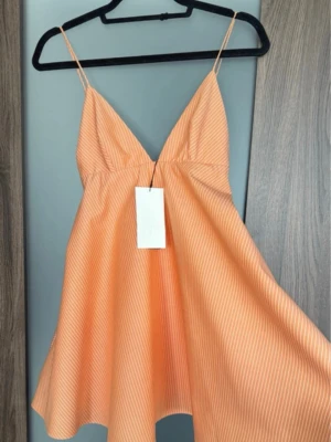  Ny trendig orange sommarklänning från Zara - Säljer en söt orange klänning från Zara med tunna vita ränder. Klänningen har smala axelband, djup V-ringning och en utställd, luftig passform. Perfekt för varma dagar och ger en fräsch och trendig look.