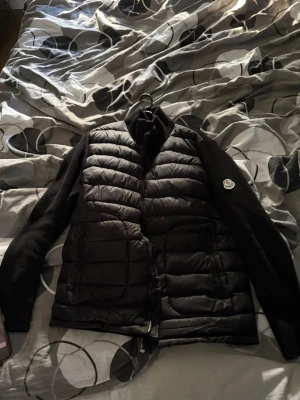 Svart dunjacka från Moncler - Snygg svart dunjacka från Moncler med quiltad front och rygg samt stickade armar. Jackan har hög krage, dragkedja framtill och Moncler-logga på ena armen. Perfekt för kalla vinterdagar och riktigt trendig stil.