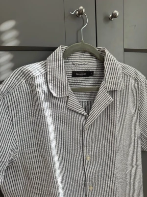 Randig linneskjorta från Dressmann - Kortärmad skjorta från Dressmann i tunn och luftig linnemix. Skjortan har smala svarta och vita ränder, klassisk krage och knäppning framtill. Perfekt för varma dagar och enkel att styla med jeans eller shorts. Helt ny