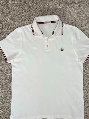 Vit Moncler pikétröja med röd detalj - Vit pikétröja från Moncler med klassisk krage och röda samt svarta ränder på krage och ärmslut. Tröjan har korta ärmar, knappar framtill och Moncler-logga på bröstet. Perfekt för en clean och sportig look.