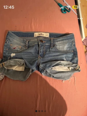 Blå lowwast jeansshorts från Hollister - Säljer ett par blå jeansshorts från Hollister med slitna detaljer och upprullade benslut. Klassisk femficksmodell med knapp och dragkedja framtill. Perfekta till sommaren och har den ikoniska Hollister-loggan på fickan. Gjorda i mjuk denim för skön känsla. Lägger ut igen för gratis frakt 